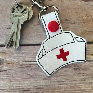 May include: White nurse's hat keychain with a red cross and a red button. The keychain is attached to a silver key ring.