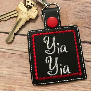 May include: Black leather keychain with a red button and white embroidery that says "Yia Yia" in a cursive font.