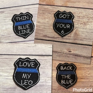 May include: Four embroidered patches with a blue line and white text on a black background. The patches are shaped like a police badge. The text on the patches reads "Thin Blue Line", "Got Your 6", "Love My Leo", and "Back The Blue".