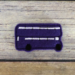 May include: A purple felt double-decker bus with white window details and black wheels. The bus is a handmade craft item, likely a patch or applique, and is set against a wooden background.