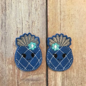 Op de afbeelding: Twee blauwe vilt ananas-applicaties met geborduurde details. Elke ananas heeft een zilveren en blauwe kroon, zwarte ogen en een witte glimlach.