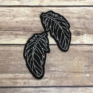 以下が含まれることがあります： 白い刺繍の羽根模様が施された黒いフェルト生地の2つのパッチ