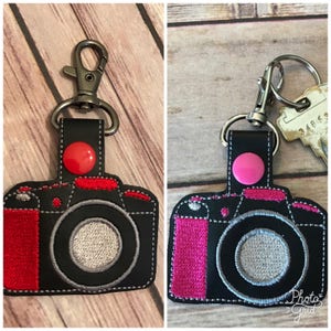 May include: Two black camera-shaped keychains with red and pink accents. The keychains have a silver metal loop and a button on the top.