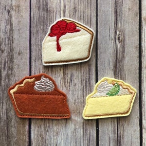 Puede incluir: Tres rebanadas de pastel de fieltro con detalles bordados. La primera rebanada es una tarta de cerezas con una cereza roja en la parte superior. La segunda rebanada es una tarta de chocolate con un remolino de glaseado blanco. La tercera rebanada es una tarta de lima con una rodaja de lima verde en la parte superior.