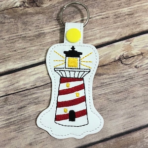 May include: White leather keychain with a red and white striped lighthouse embroidered on it. The lighthouse has a yellow light at the top and a black door.