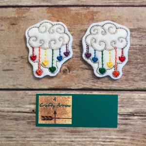 May include: Two white felt cloud appliques with rainbow colored strings hanging down from them. Each string has a small red, orange, yellow, green, blue, indigo, and violet heart attached to it. The text "A Crafty Arrow" is visible in the bottom center of the image.
