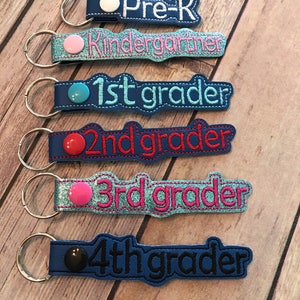 May include: Six blue keychains with different coloured buttons and embroidered text. The text reads "Pre-K", "Kindergarten", "1st grader", "2nd grader", "3rd grader", and "4th grader".