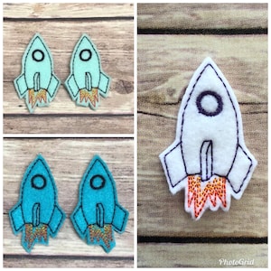 May include: Three felt rocket ship appliques. One is white with purple trim, one is light blue with orange trim, and one is turquoise with orange trim.