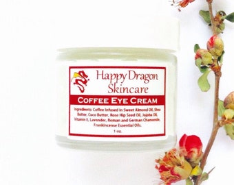 May include: A small white jar with a red label that reads "Happy Dragon Skincare" and "Coffee Eye Cream". The jar is sitting on a white background with a sprig of pink flowers.