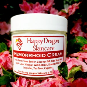 May include: A white jar with a red label that reads "Happy Dragon Skincare" and "Hemorrhoid Cream". The label lists ingredients including shea butter, coconut oil, aloe vera gel, apple cider vinegar, witch hazel, and essential oils of lavender, tea tree, and cypress. The jar is sitting on a bed of pink flower petals.