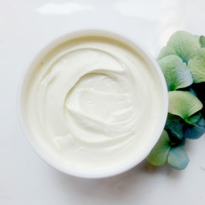 K&ouml;nnte beinhalten: Draufsicht auf ein wei&szlig;es, cremiges Produkt in einem runden Beh&auml;lter. Das Produkt hat eine glatte, gewirbelte Textur. Ein Strau&szlig; blauer und gr&uuml;ner Hortensienbl&uuml;ten befindet sich im Hintergrund und verleiht der Szene einen Hauch von nat&uuml;rlicher Sch&ouml;nheit.