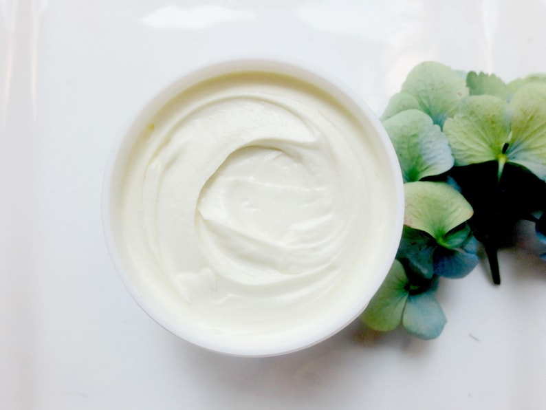 K&ouml;nnte beinhalten: Draufsicht auf ein wei&szlig;es, cremiges Produkt in einem runden Beh&auml;lter. Das Produkt hat eine glatte, gewirbelte Textur. Ein Strau&szlig; blauer und gr&uuml;ner Hortensienbl&uuml;ten befindet sich im Hintergrund und verleiht der Szene einen Hauch von nat&uuml;rlicher Sch&ouml;nheit.