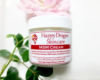 May include: A white jar with a red label that says "Happy Dragon Skincare" and "MSM CREAM". The label also lists ingredients, directions, and a website address. The jar is sitting on a white surface with a pink rose and green leaves.