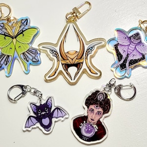 May include: Five iridescent keychains with different designs.  A green and yellow moth with crystals, a gold helmet, a purple moth with stars, a purple bat, and a woman with a crystal ball.