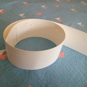 May include: A white plastic punch tape with holes for a computer or other machine. The tape is rolled up and has a flat section extending from the roll.