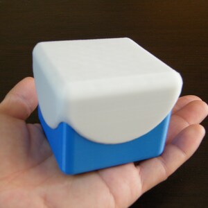 May include: A small, white box with a blue base. The box has a rounded top and a smooth, matte finish.