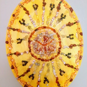May include: A circular sun-shaped mosaic made of small stones in shades of yellow, orange, red, and brown. The mosaic is set against a yellow background with a red and orange border.