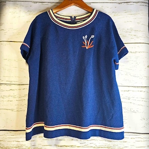 May include: A short-sleeved blue knit top featuring a round neckline with cream and red striped trim. The hem and sleeve cuffs also have matching striped detailing. A small embroidered plant motif in white and orange sits on the upper left chest.