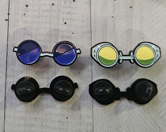 May include: Two pairs of enamel pins in the shape of goggles. One pair has blue lenses, the other has yellow and green lenses. The pins have black backs with a butterfly clutch.