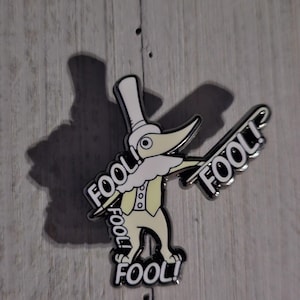May include: A metal enamel pin featuring a pale yellow cartoon character wearing a tall top hat and a vest. The character has a long pointed nose and is surrounded by the word FOOL! printed in bold white letters with black outlines.