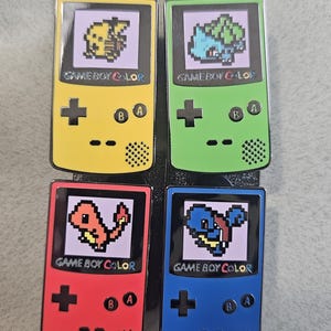 May include: Four enamel pins featuring the Game Boy Color handheld gaming console with different Pokemon characters on the screen. The pins are yellow, green, red, and blue.
