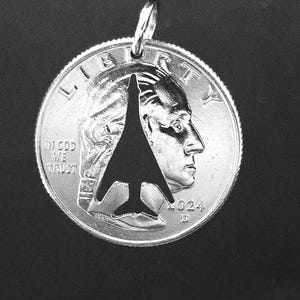 May include: A silver coin pendant with a cut-out jet aircraft silhouette. The coin is a quarter with George Washington's profile. The pendant is attached to a silver loop. The year 2024 is visible.