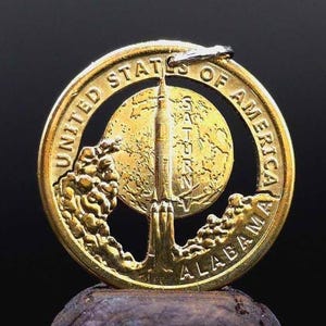 May include: A gold-colored coin featuring a rocket launching into space, with the words "UNITED STATES OF AMERICA" and "ALABAMA" inscribed around the edges. The coin also has the word "SATURN" and an image of the Earth.