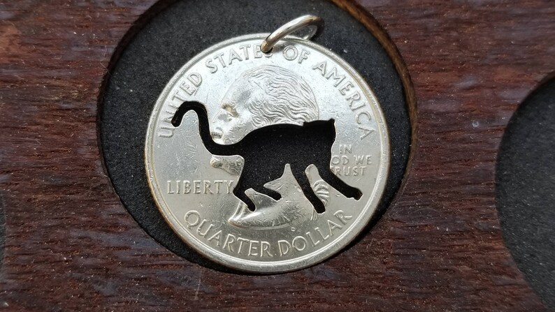 Może przedstawiać: Srebrna amerykańska moneta o wartości ćwierć dolara z wyciętym czarnym siluetem pantery. Na monecie widnieją napisy "Liberty", "In God We Trust" i "Quarter Dollar".
