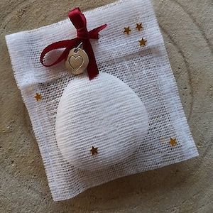 Peut inclure: Une boule ronde blanche recouverte de tissu avec un charme en forme de cœur argenté attaché à un ruban rouge. L'ornement est posé sur un tissu blanc en maille avec des étoiles dorées.