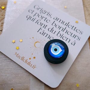 Peut inclure: Un porte-bonheur rond noir avec un motif d'œil bleu et blanc, posé sur une carte avec du texte en français et des étoiles dorées. La carte est sur une surface claire, avec le site Mzellelulu.fr imprimé.