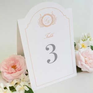 May include: White table number card with a gold border and a gold monogram in the top center. The card reads "Table 3" in gray text.