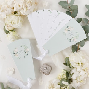 Puede incluir: Dos abanicos de boda de color azul claro con diseños florales blancos y las iniciales "J & T" en el centro. Los abanicos están decorados con flores blancas y hojas verdes. Los abanicos están atados con una cinta blanca.