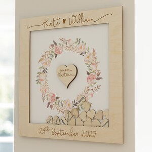 Puede incluir: Un marco de madera con un dise&ntilde;o de corona de flores y un recorte en forma de coraz&oacute;n en el centro. El marco est&aacute; personalizado con los nombres "Kate & William" y la fecha "24 de septiembre de 2027".