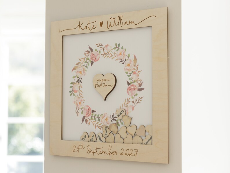 Puede incluir: Un marco de madera con un dise&ntilde;o de corona de flores y un recorte en forma de coraz&oacute;n en el centro. El marco est&aacute; personalizado con los nombres "Kate & William" y la fecha "24 de septiembre de 2027".