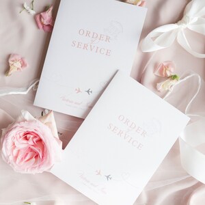 Könnte beinhalten: Zwei weiße Trauungsprogramme mit rosa und grauen Akzenten. Die Programme zeigen den Text "Order of Service" und eine Grafik mit zwei Flugzeugen mit Herzen. Der Text "Victoria und Liam" ist ebenfalls enthalten.