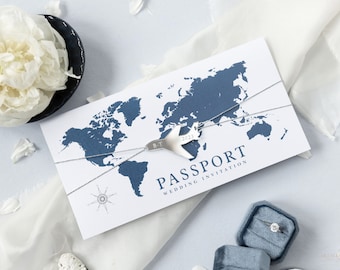May include: A white wedding invitation with a blue world map and a silver airplane charm. The invitation says "Passport Wedding Invitation 2023" and has a compass design.
