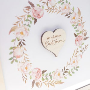 Puede incluir: Un marco blanco con una corona de flores de acuarela y un coraz&oacute;n de madera con el texto "Mr & Mrs Bertram" en el centro.
