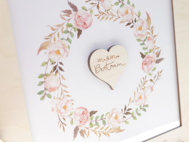 Puede incluir: Un marco blanco con una corona de flores de acuarela y un coraz&oacute;n de madera con el texto "Mr & Mrs Bertram" en el centro.