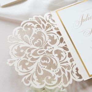 May include: A white wedding invitation with a delicate, intricate, laser-cut design. The invitation features a floral pattern with a gold border.