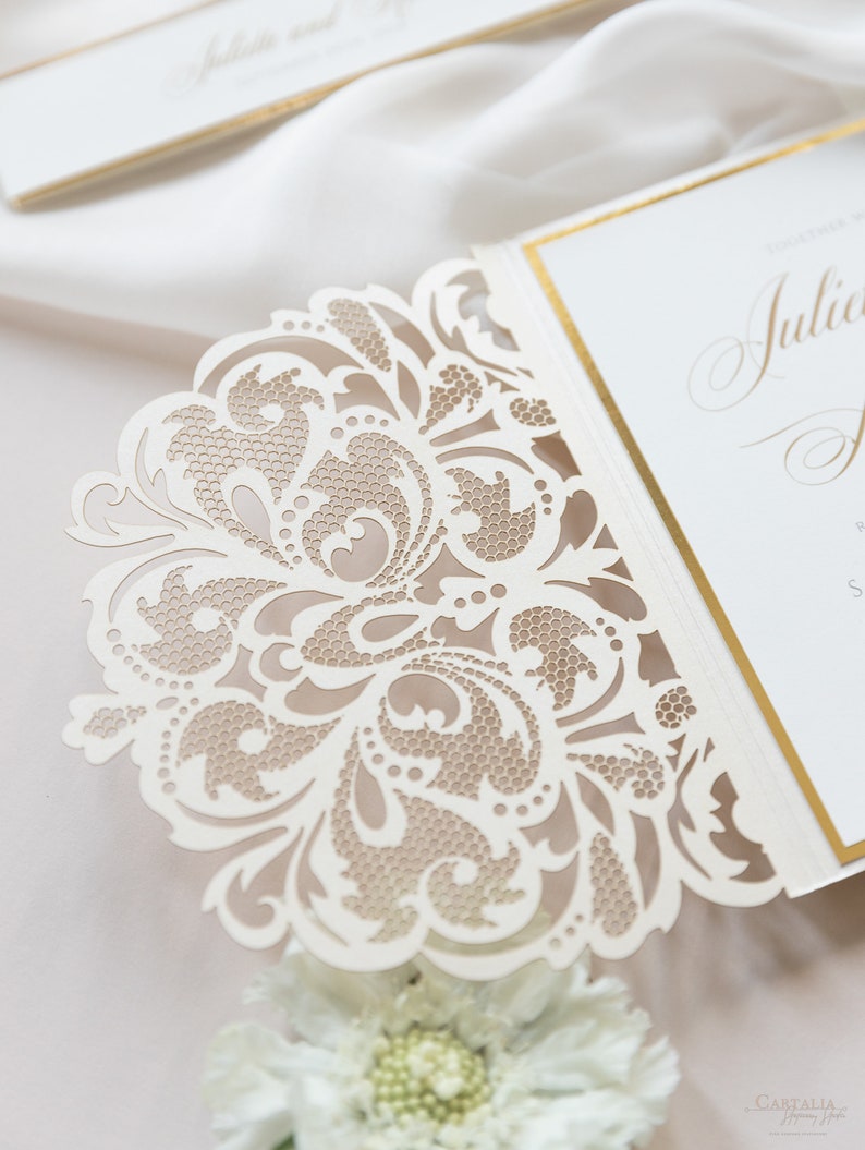 May include: A white wedding invitation with a delicate, intricate, laser-cut design. The invitation features a floral pattern with a gold border.