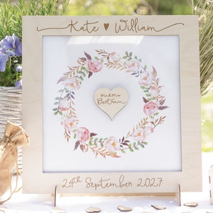 Puede incluir: Un marco de madera para libro de visitas de boda con un dise&ntilde;o de corona de flores. El marco tiene un recorte en forma de coraz&oacute;n en el centro con el texto "m&m Bertram" en su interior. El marco est&aacute; grabado con los nombres "Kate William" y la fecha "24 de septiembre de 2027".