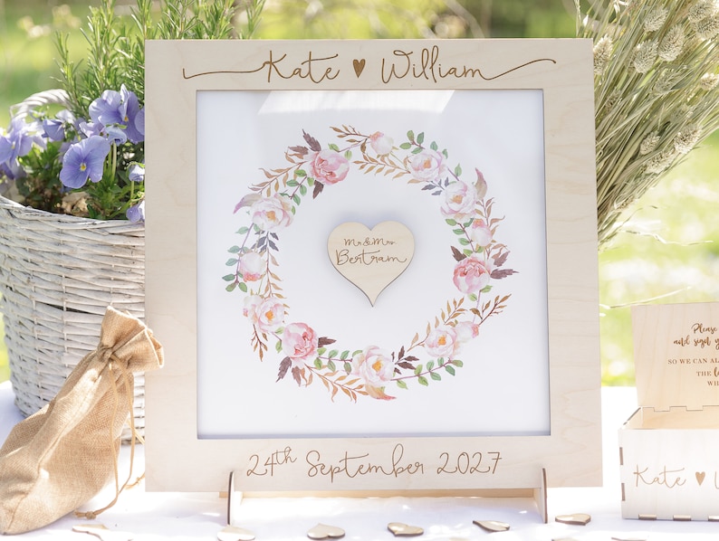 Puede incluir: Un marco de madera para libro de visitas de boda con un dise&ntilde;o de corona de flores. El marco tiene un recorte en forma de coraz&oacute;n en el centro con el texto "m&m Bertram" en su interior. El marco est&aacute; grabado con los nombres "Kate William" y la fecha "24 de septiembre de 2027".