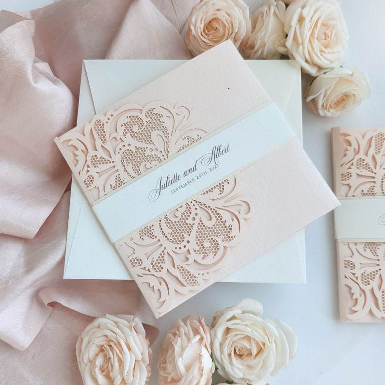 May include: A blush pink wedding invitation with a delicate laser-cut floral design. The invitation reads "Juliette and Albert, September 24th, 2021".