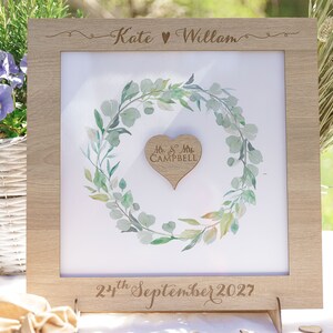 Puede incluir: Un marco de madera con fondo blanco y una ilustraci&oacute;n de acuarela de una corona de eucalipto verde. La corona rodea un coraz&oacute;n de madera con el texto "Mr & Mrs Campbell" grabado en &eacute;l. El marco tiene el texto "Kate &hearts; William" y "22 de septiembre de 2027" grabado en &eacute;l.