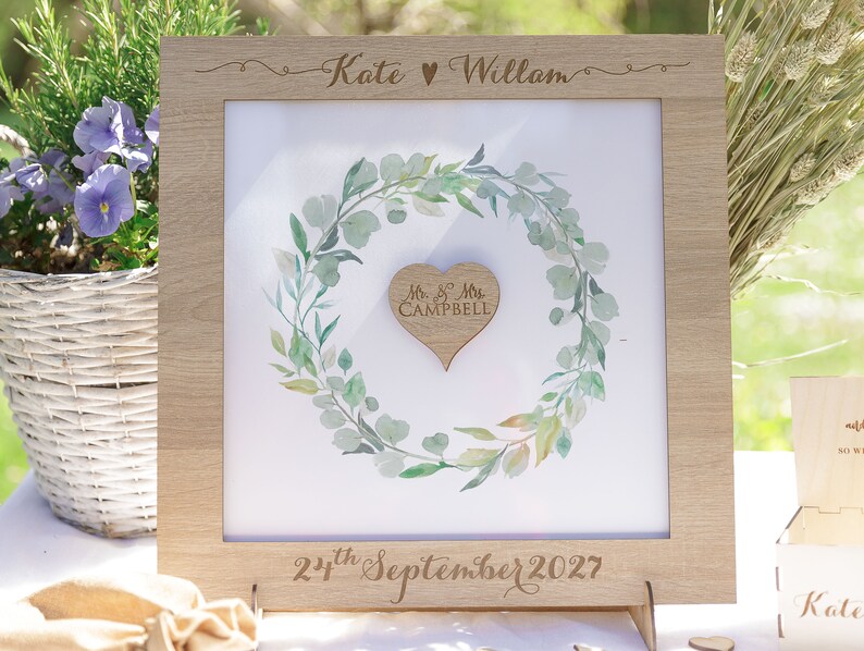 Puede incluir: Un marco de madera con fondo blanco y una ilustraci&oacute;n de acuarela de una corona de eucalipto verde. La corona rodea un coraz&oacute;n de madera con el texto "Mr & Mrs Campbell" grabado en &eacute;l. El marco tiene el texto "Kate &hearts; William" y "22 de septiembre de 2027" grabado en &eacute;l.