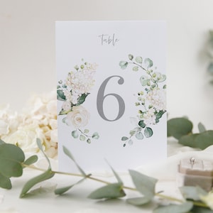 May include: White floral table number card with the number 6 in grey text and the word "Table" above the number. The card is surrounded by white flowers and green leaves.