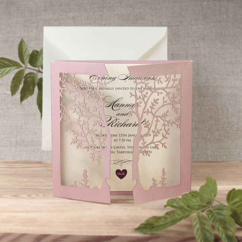 May include: Pink laser cut wedding invitation with a tree design. The invitation reads "You are cordially invited to the wedding of Hannah and Richard" and includes the date and time of the wedding. The invitation has a small heart with the initials "H&R" in the center.