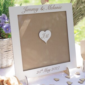Puede incluir: Marco de madera blanco con fondo marr&oacute;n y un recorte en forma de coraz&oacute;n con las iniciales "JM" en su interior. El marco est&aacute; sobre una mesa con un cartel que dice "Jimmy & Melanie" y "25 de mayo de 2027".