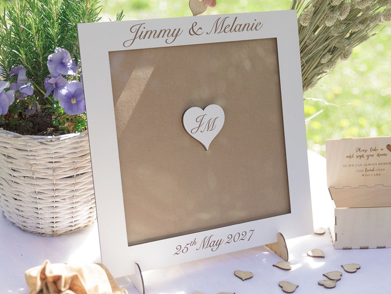 Puede incluir: Marco de madera blanco con fondo marr&oacute;n y un recorte en forma de coraz&oacute;n con las iniciales "JM" en su interior. El marco est&aacute; sobre una mesa con un cartel que dice "Jimmy & Melanie" y "25 de mayo de 2027".