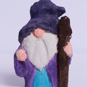 May include: A wooden figurine of a wizard with a purple robe, a blue hat, and a white beard. The wizard is holding a brown staff.